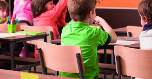 Children in elementary school are raised hand in clasroom. Teacher is in the background in front of chalkboard.